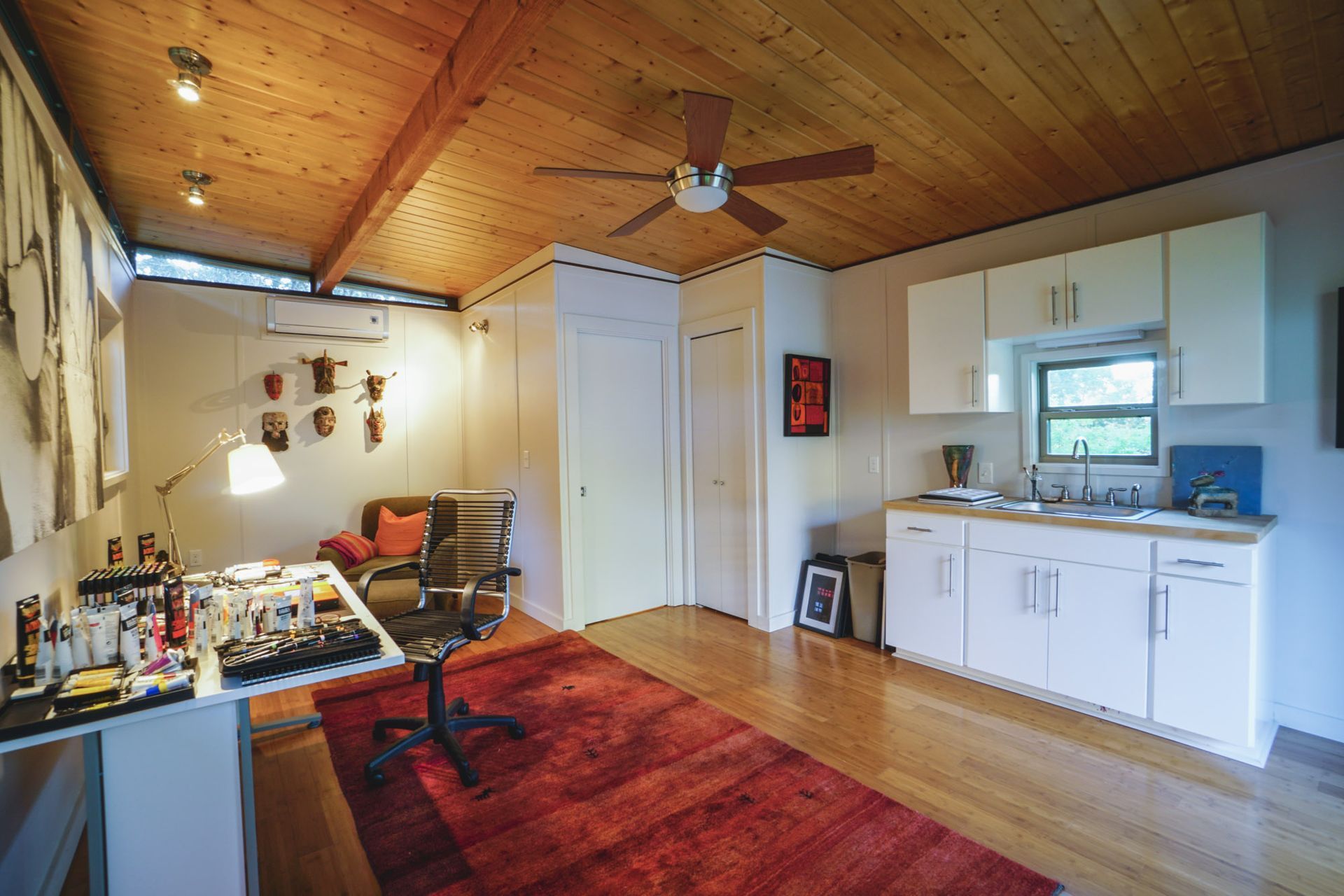 Cozy art studio with red rug, wooden ceiling, and white cabinets. Workspace with desk, chair, and art supplies.