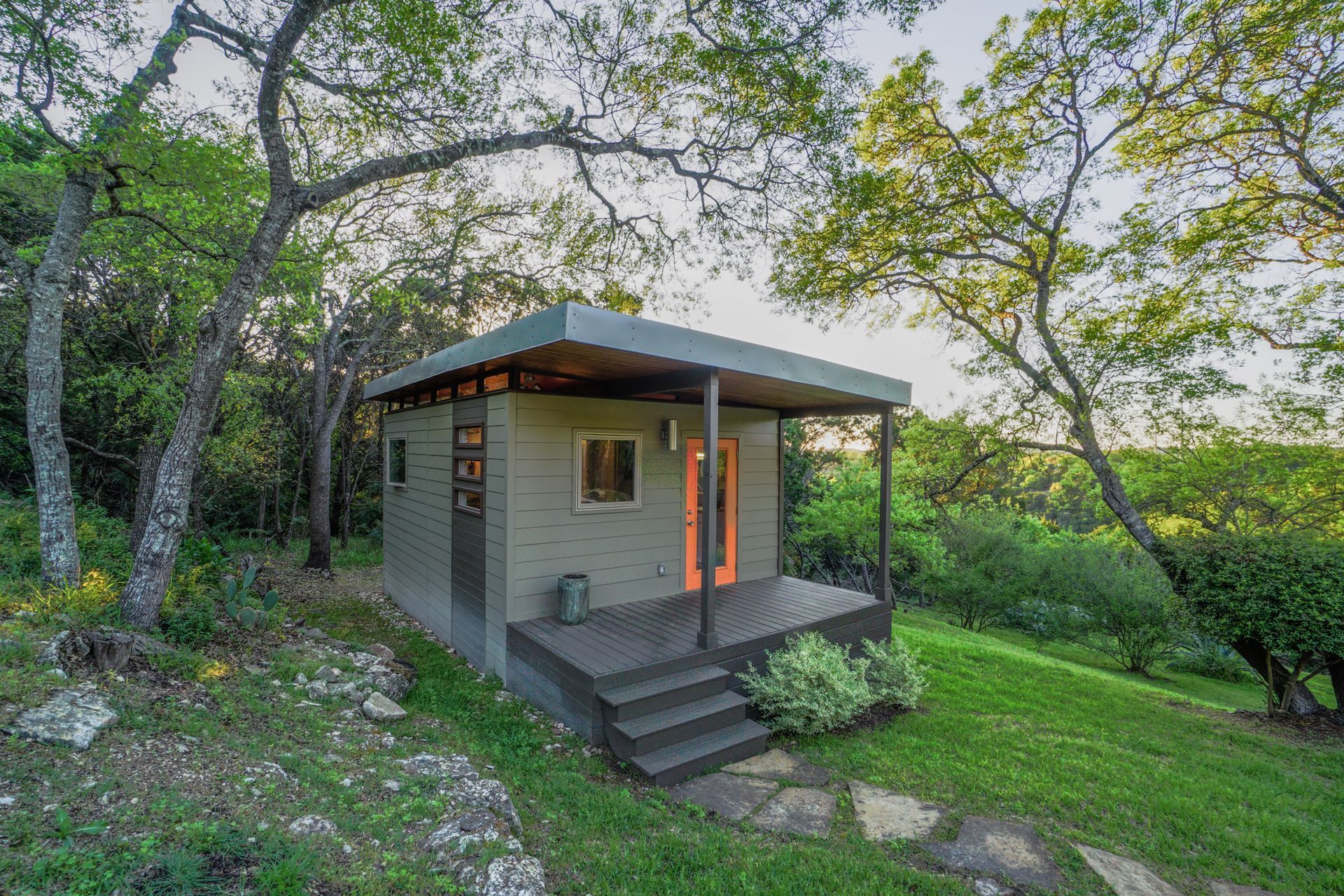 Small gray cabin with orange double doors, front porch, and steps set in a grassy, wooded area.