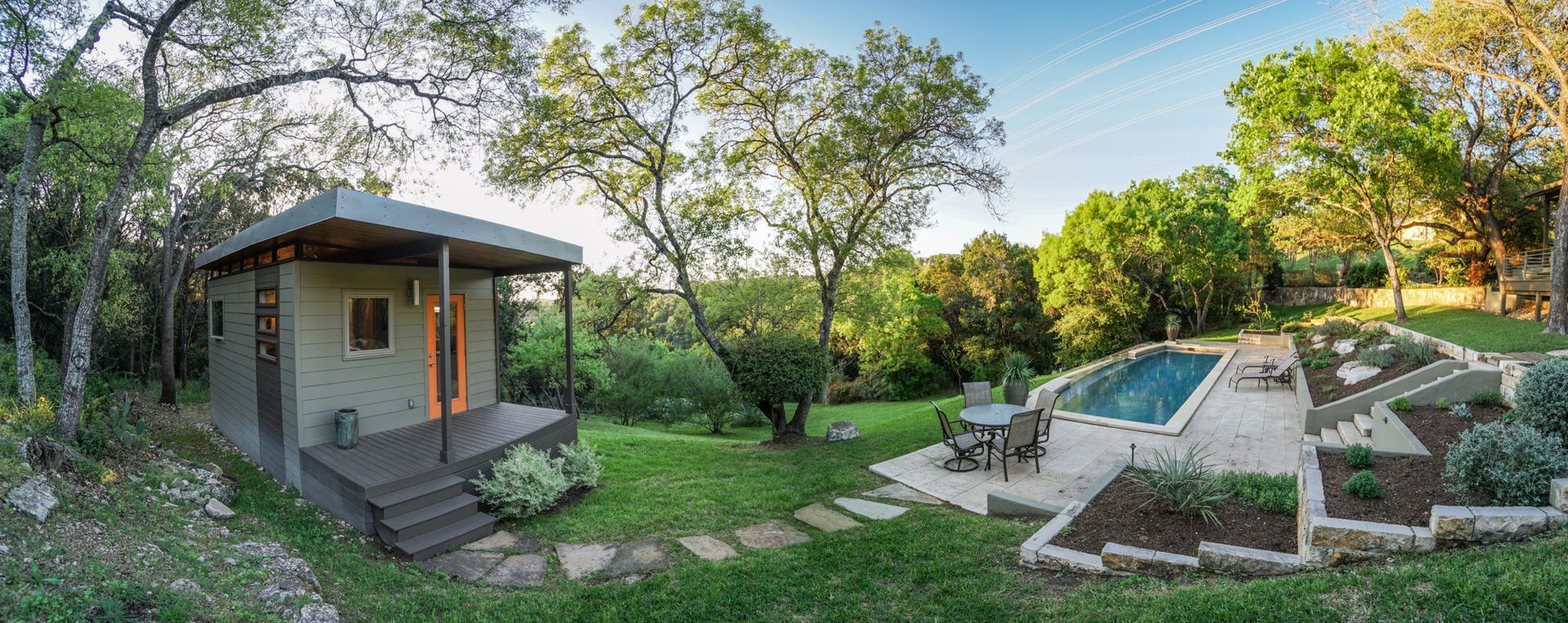A small cabin with a porch and steps is surrounded by trees and a lawn with a pool.