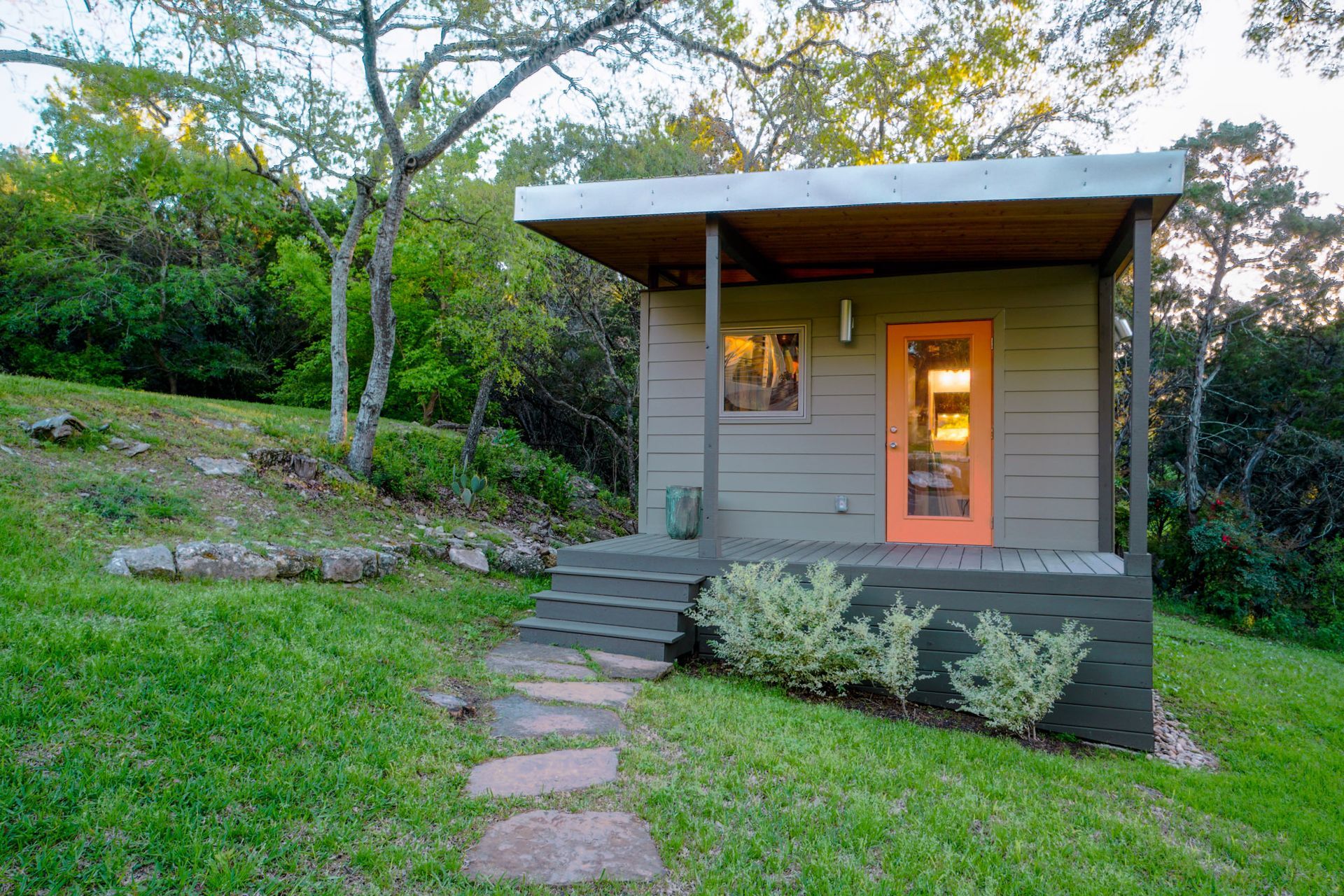 Small, gray cabin with orange door on a grassy hill; stone path leads to front steps under a porch.