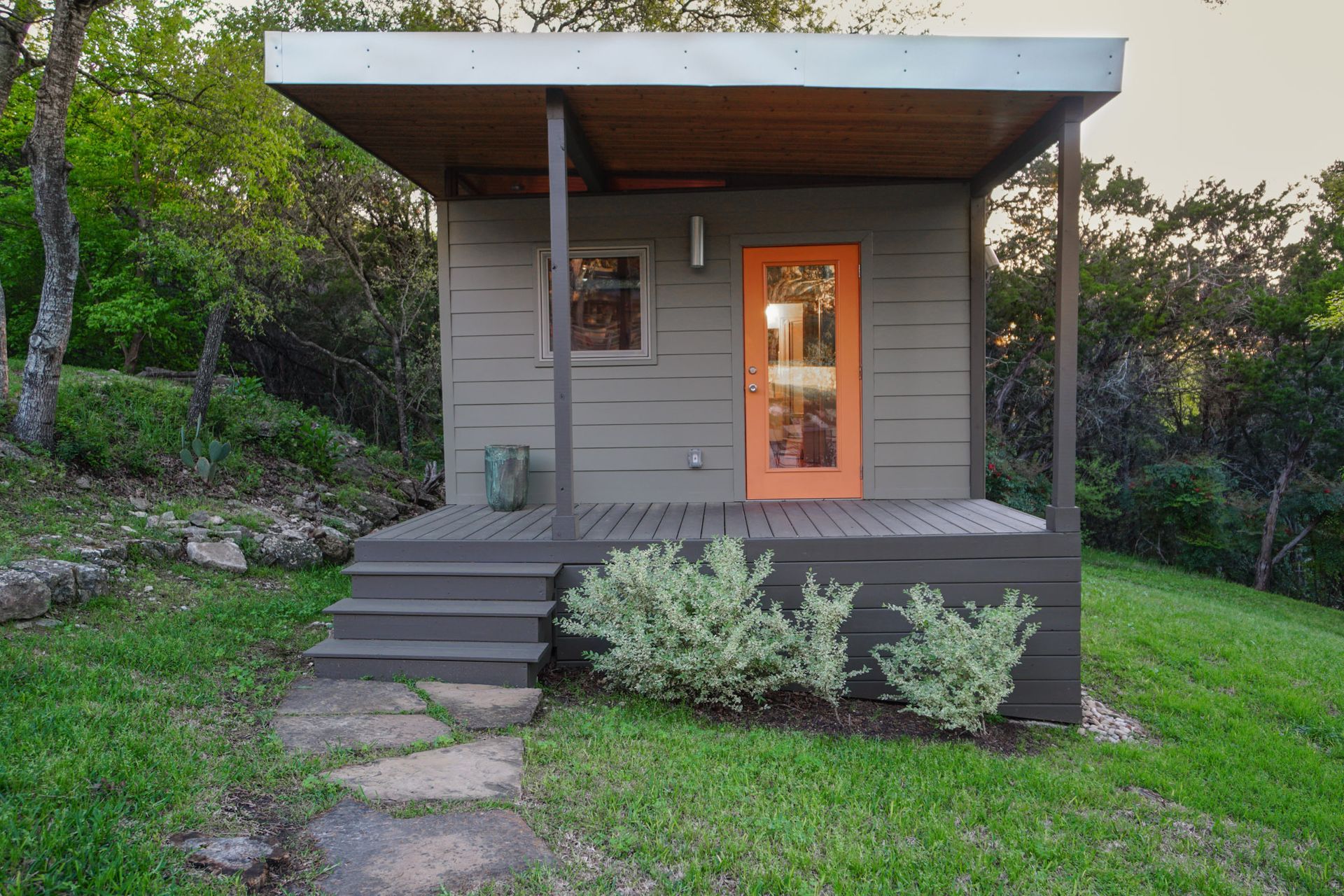 Small, gray cabin with orange door, porch, and steps on a grassy hill.