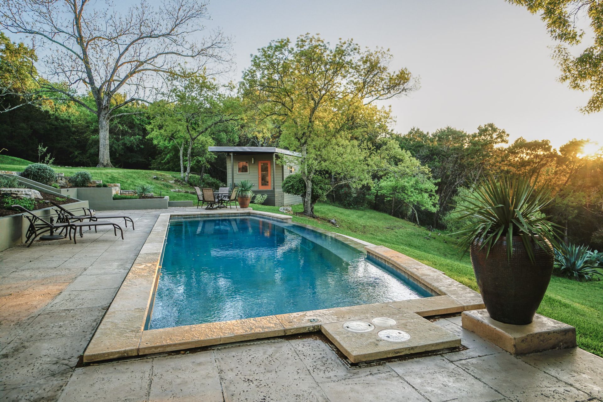 A backyard pool with a small cabana, surrounded by lush greenery, at sunset.
