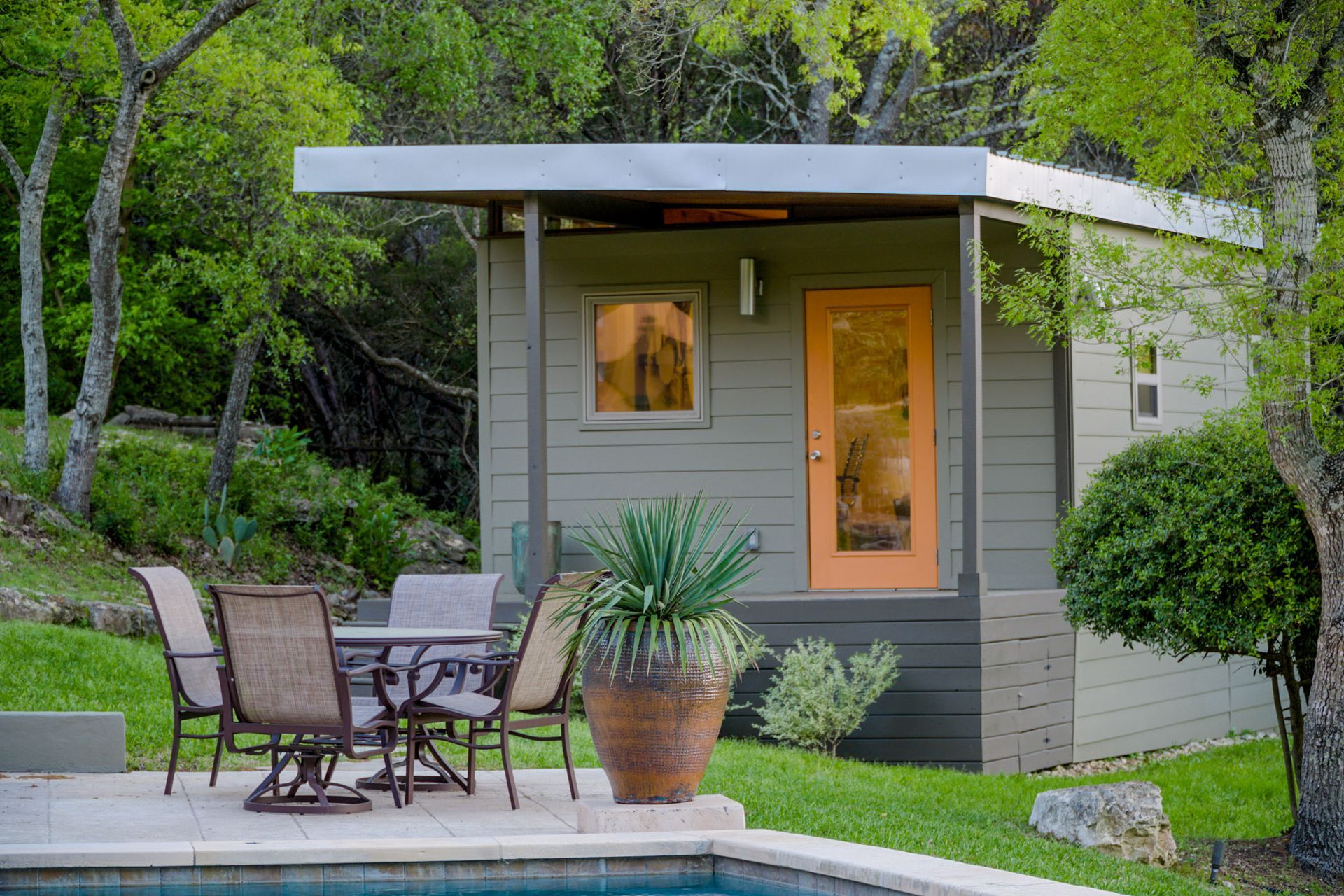 Small cabin with orange door, patio furniture by a pool, surrounded by greenery.