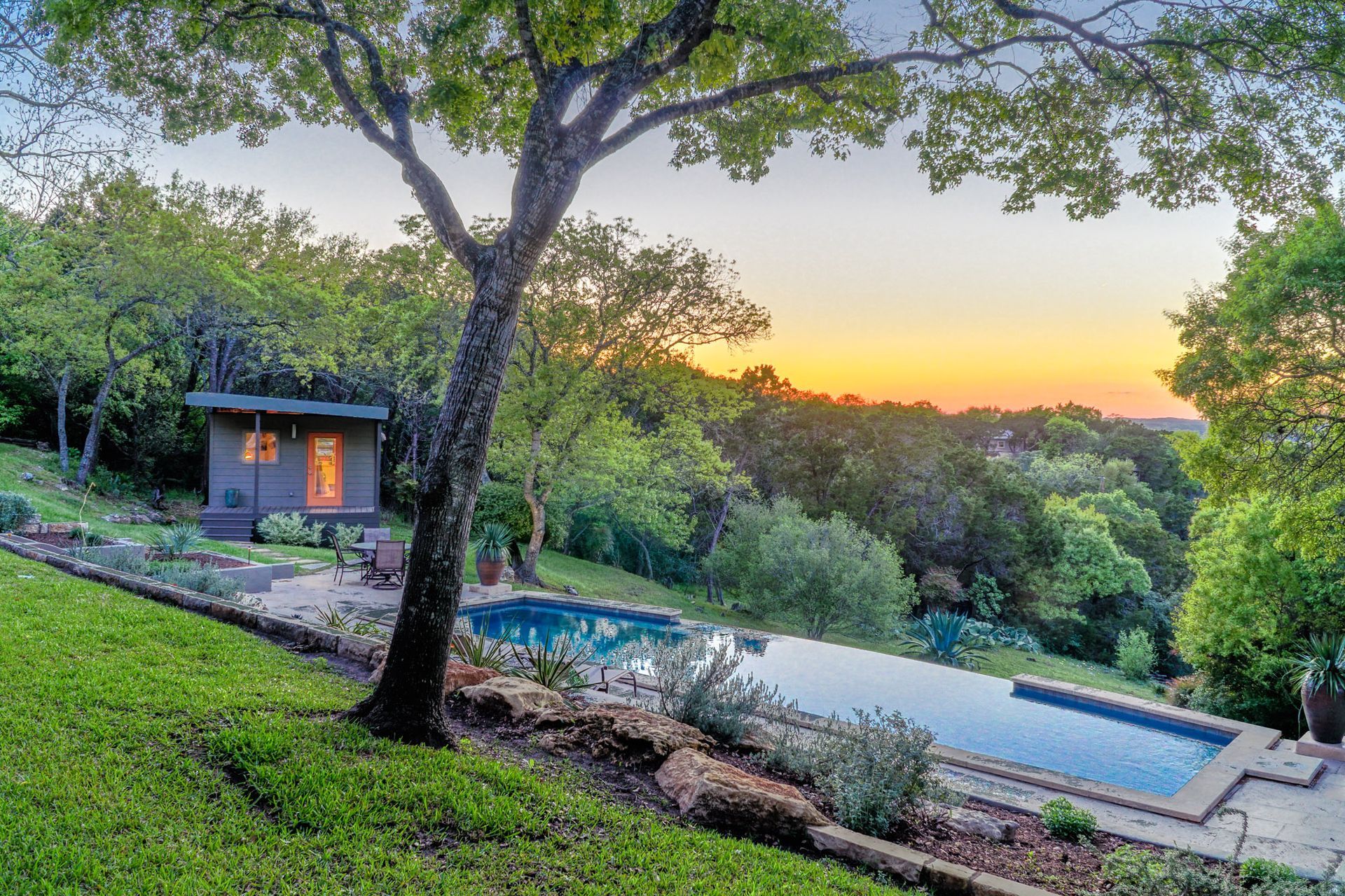 Long, rectangular pool, small wooden building, tree, and lawn on a hillside at sunset.