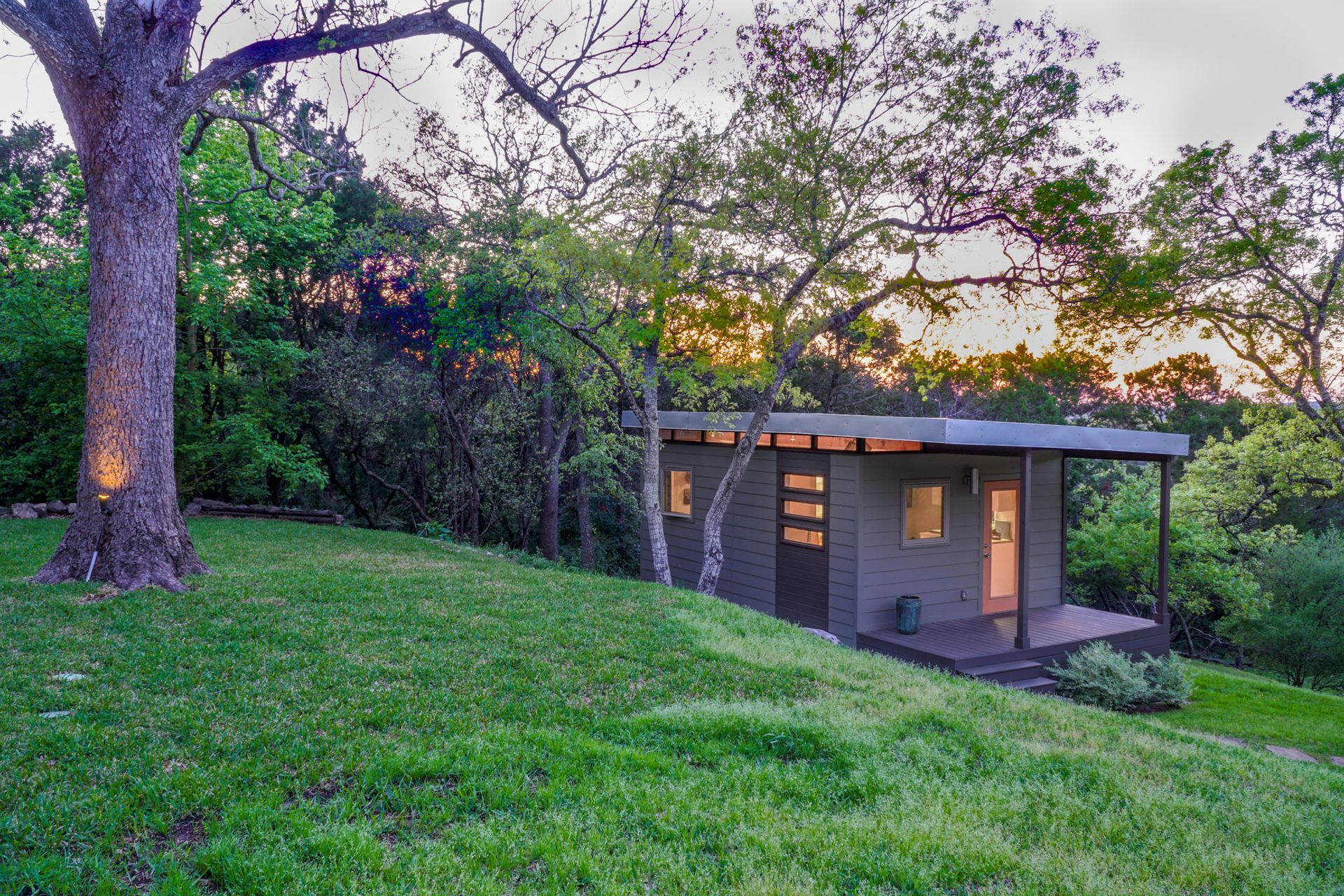 Small modern cabin on a grassy hill, surrounded by trees. Dusk lighting.