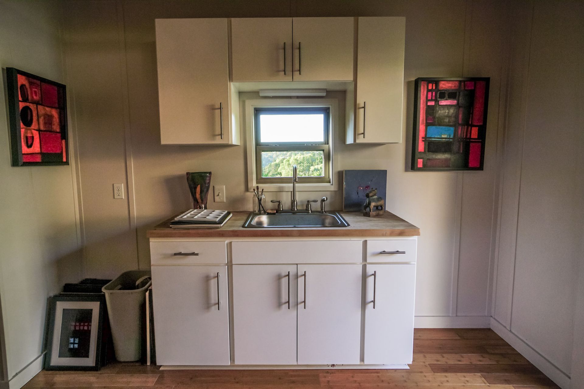 Small kitchen with cabinets, sink, and window. Art on walls.