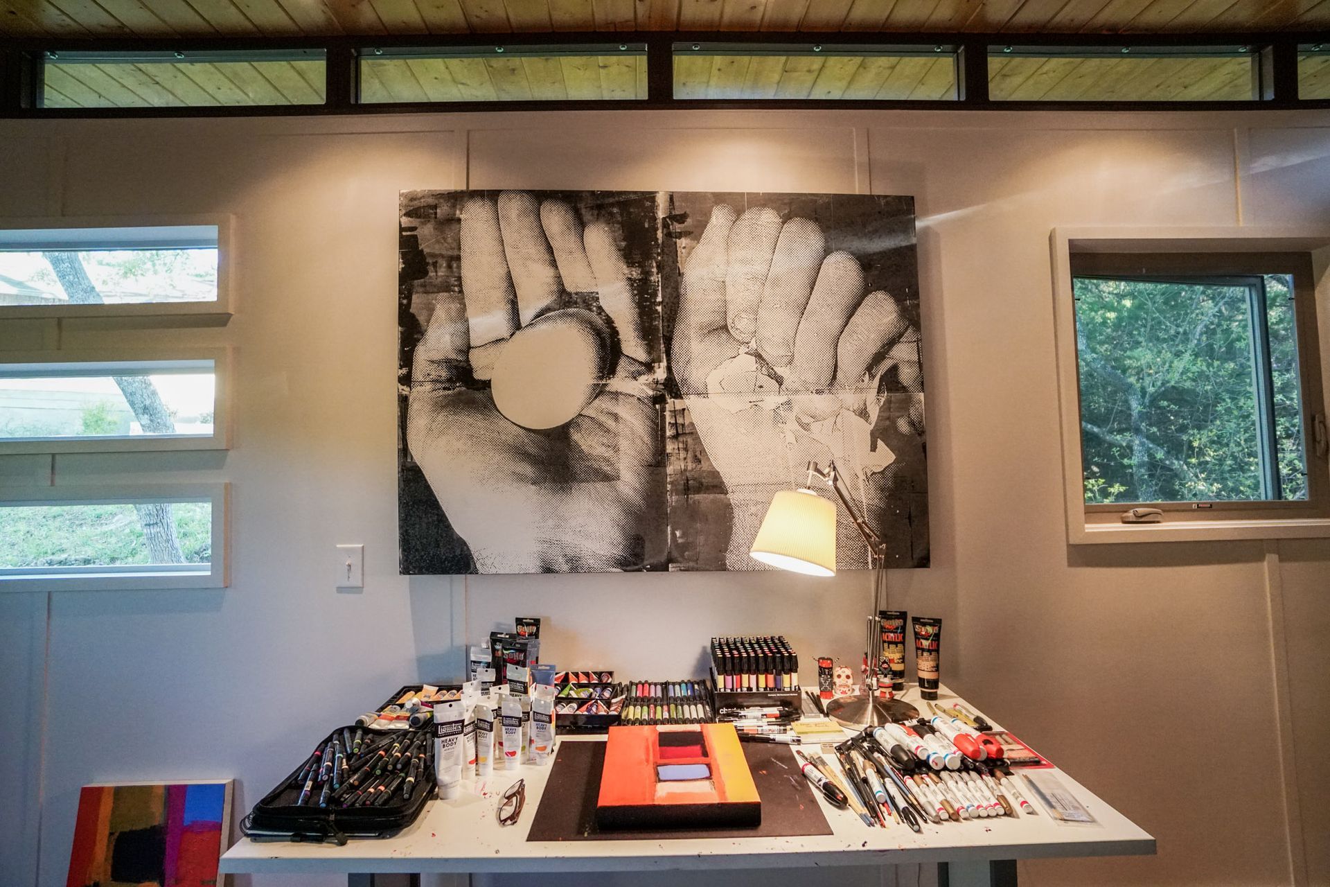 Art studio with paint supplies on a table under a two-part hand print art piece. Windows on either side.