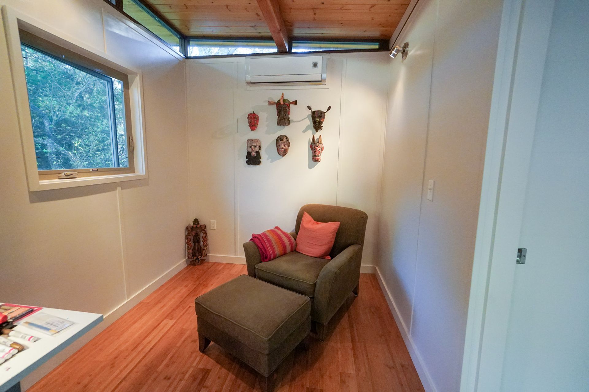 Cozy room with armchair, ottoman, masks on wall, wooden floor, and a window with a view of greenery.