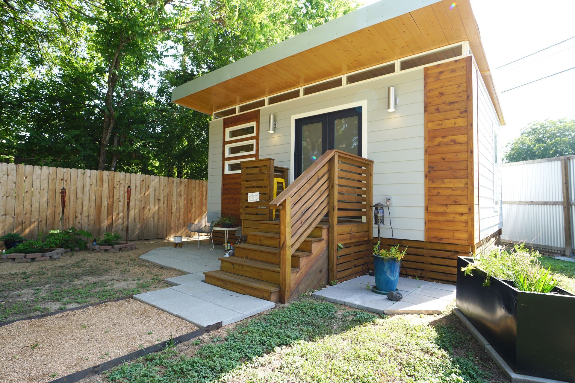 Tiny modern house with wooden accents and steps leading to a dark door. Greenery surrounds.
