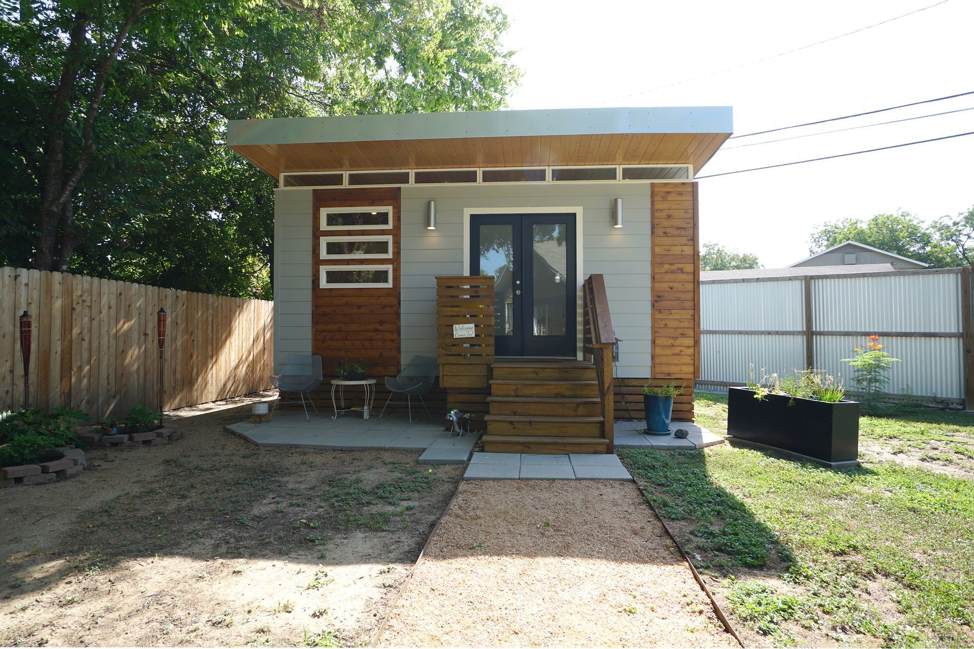 Small, modern, gray and wood-accented tiny house with a gravel path, wooden deck, and a fenced yard.