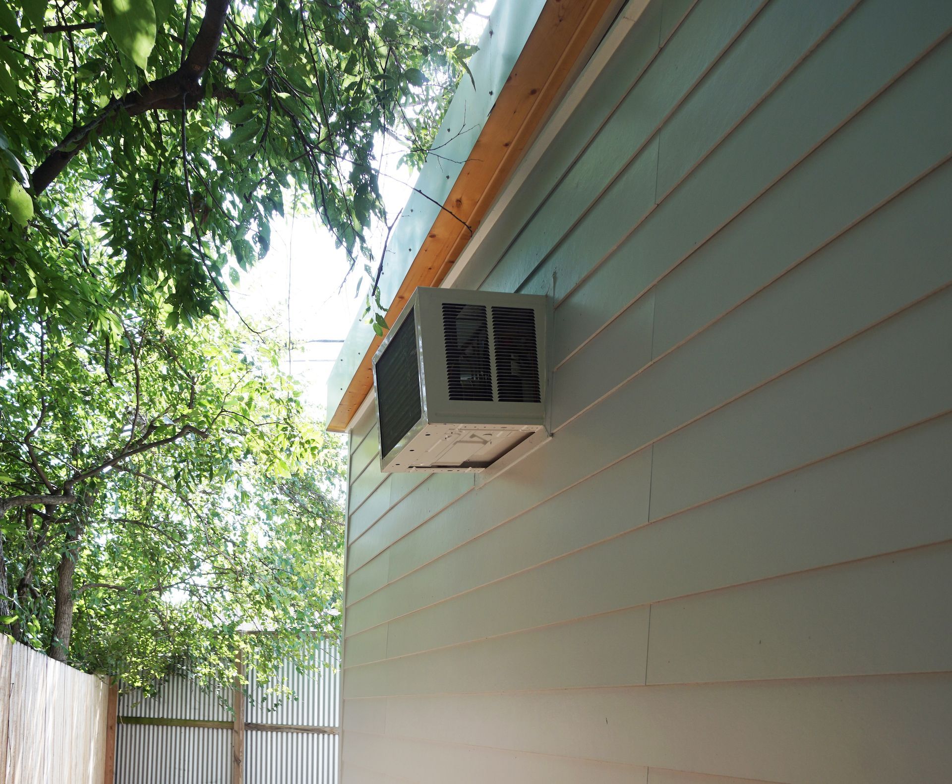 Air conditioner mounted on the light blue siding of a building near a tree and fence.