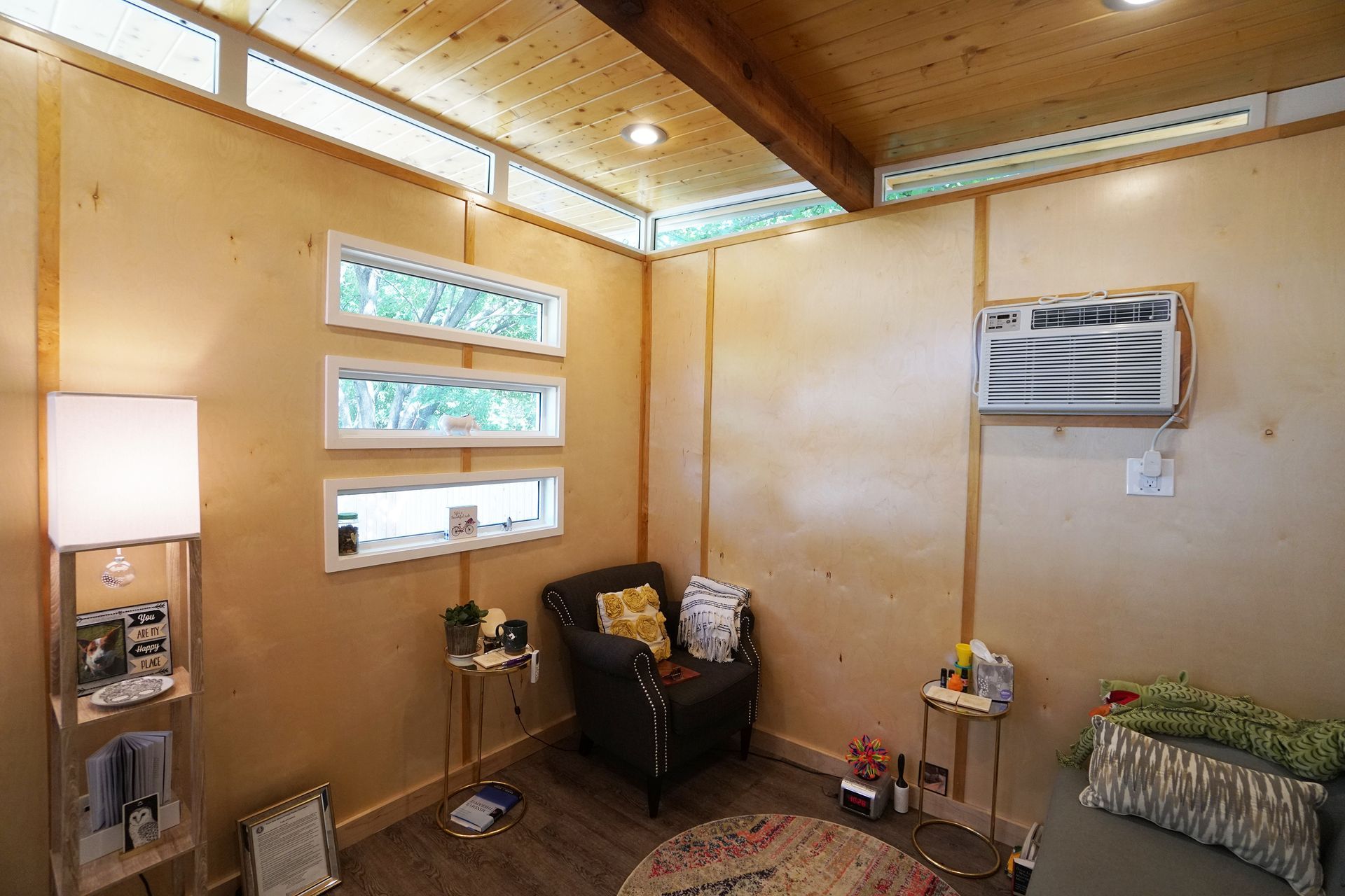 Small, light-wood-paneled room with a chair, windows, an AC unit, and a small rug.