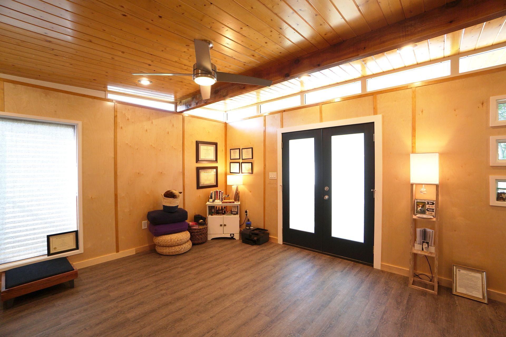 Cozy room with wood ceiling, walls, and floor. Black double doors, artwork, and a ceiling fan are visible.