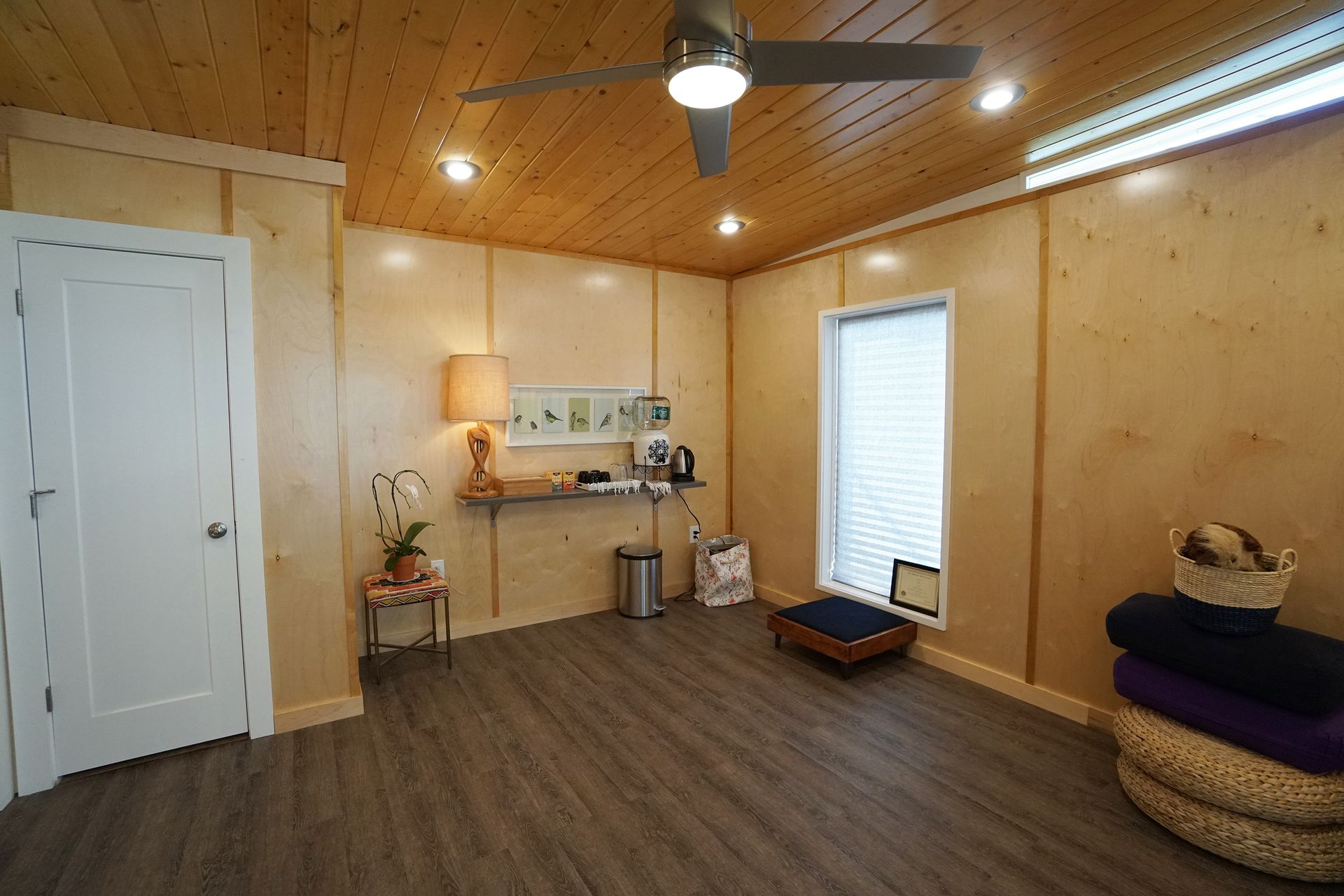 Wooden-paneled room with ceiling fan, white door, window with shades, gray flooring. Relaxation area with items.