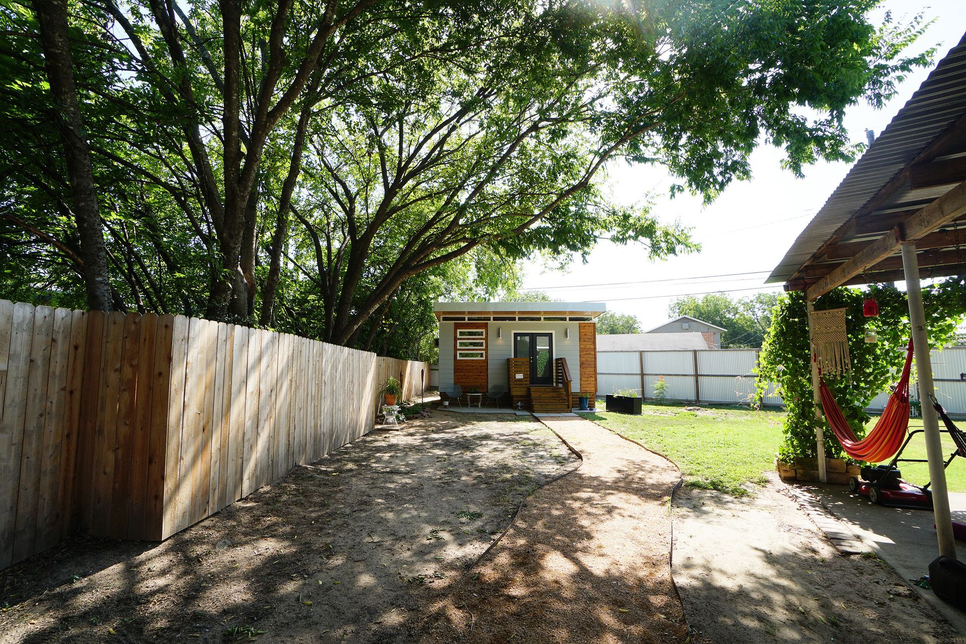 Backyard with small cabin, wooden fence, shaded area with hammock, and green lawn.