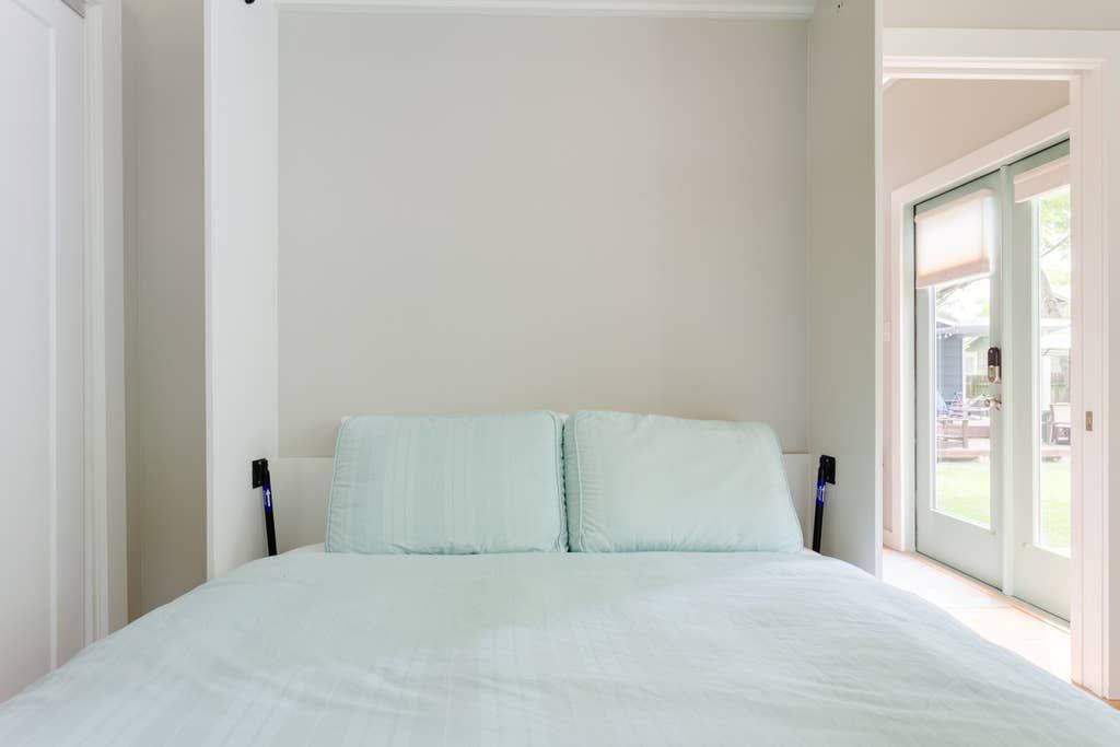 A bed with pale blue pillows and bedding, against a plain white wall, next to an open doorway.