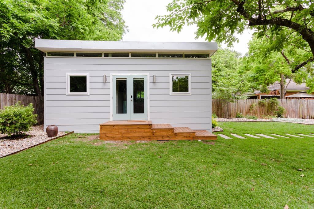 Modern grey shed with light blue doors and windows, on a green lawn with trees.