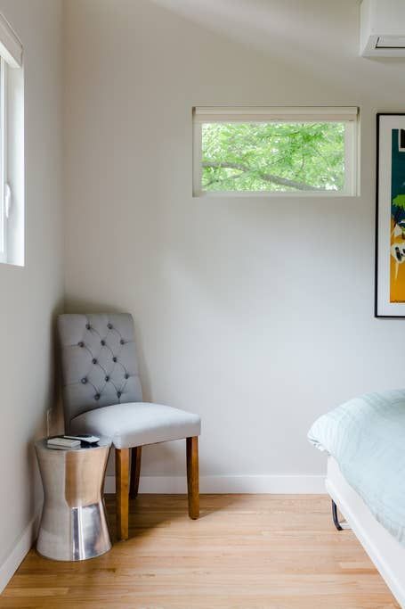 Bedroom corner with chair, stool, small window with tree view, and art. Light wood floor and white walls.