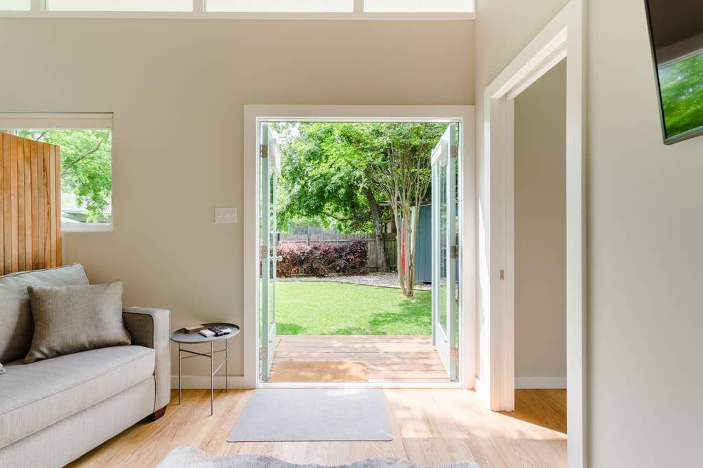 Living room with open doors to a backyard; light gray sofa, wooden floors, and a small window.