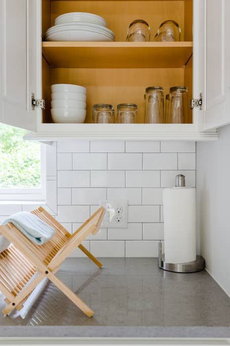 Kitchen cabinet with dishes, glasses, and paper towel roll on a gray countertop with a drying rack.
