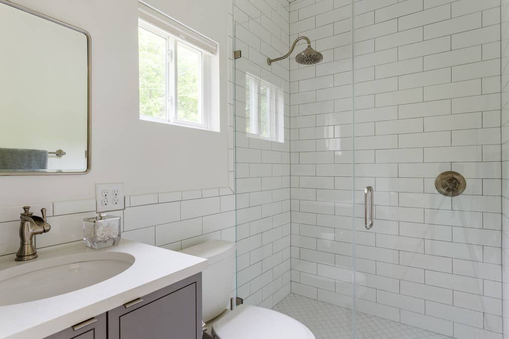 Bathroom with white brick-style tiles, gray vanity, glass shower, and bronze fixtures.