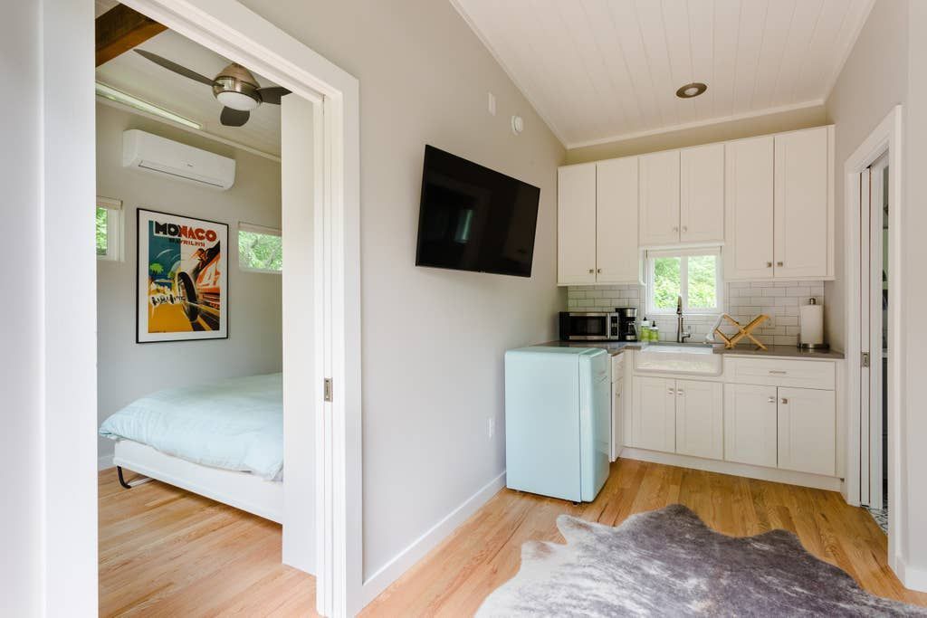 Small room with a bed, kitchen area, and TV; light blue fridge and white cabinets, wooden floor.