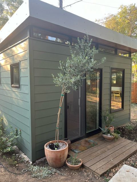 Green shed with brown trim, door, and windows. An olive tree stands in front, potted on a wood deck.
