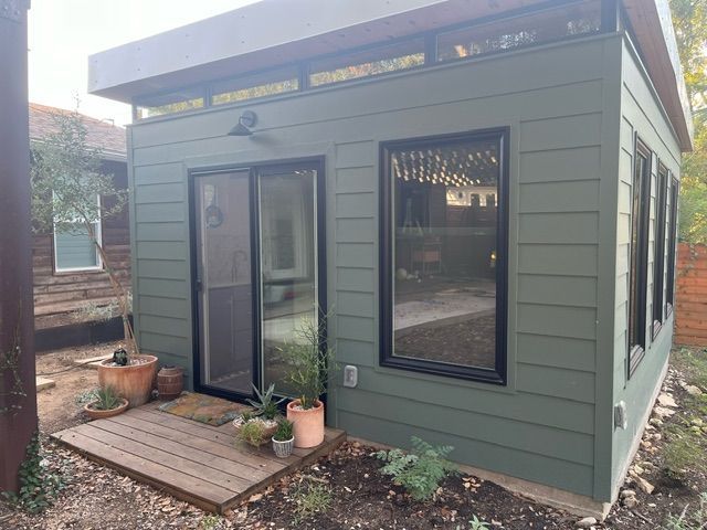 Green-sided shed with sliding glass door and windows, wooden deck, small plants, set in a yard.