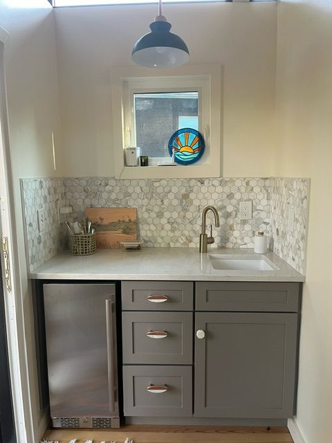 Small gray bar area with a sink, mini-fridge, cabinets, and marble backsplash. A small window is above the sink.