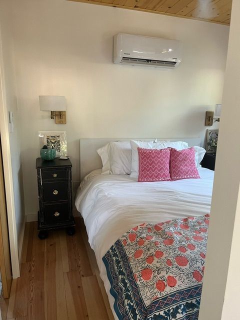 Bedroom with white walls, bed, nightstand, air conditioner, and wood flooring. Pink pillows and floral blanket on bed.