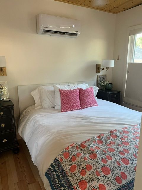 Bedroom with white bed, pink pillows, patterned throw, and wall-mounted air conditioner.