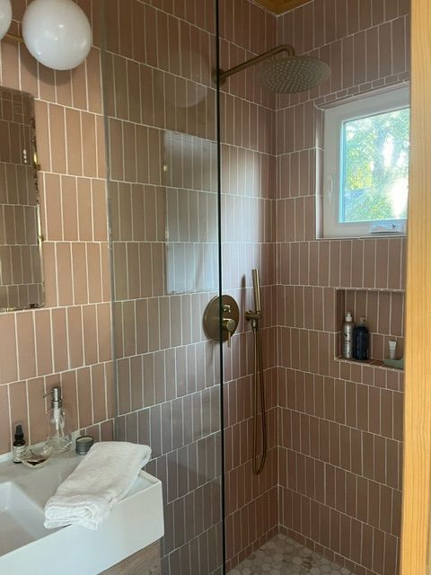 Pink tiled shower with glass door, gold fixtures, and a small window.