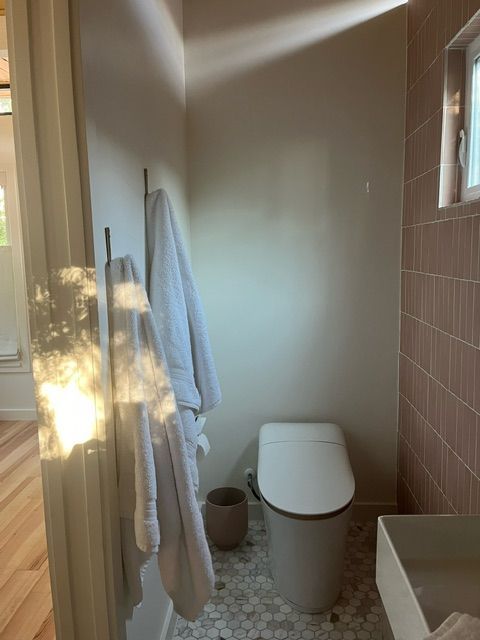 Bathroom with white toilet, pink tiled wall, towels on a rack, and a small sink. Sunlight streams in.