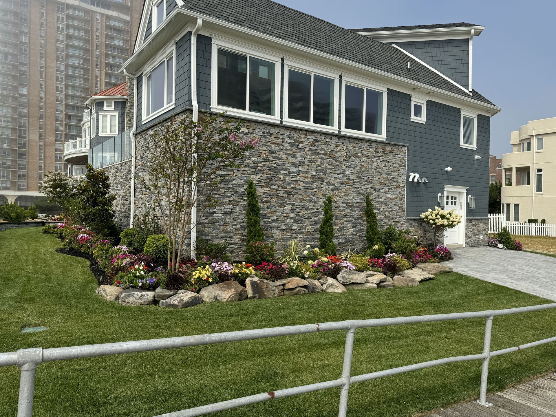 Blue-gray house with white trim and stone facade beside a flower garden and green lawn by the water