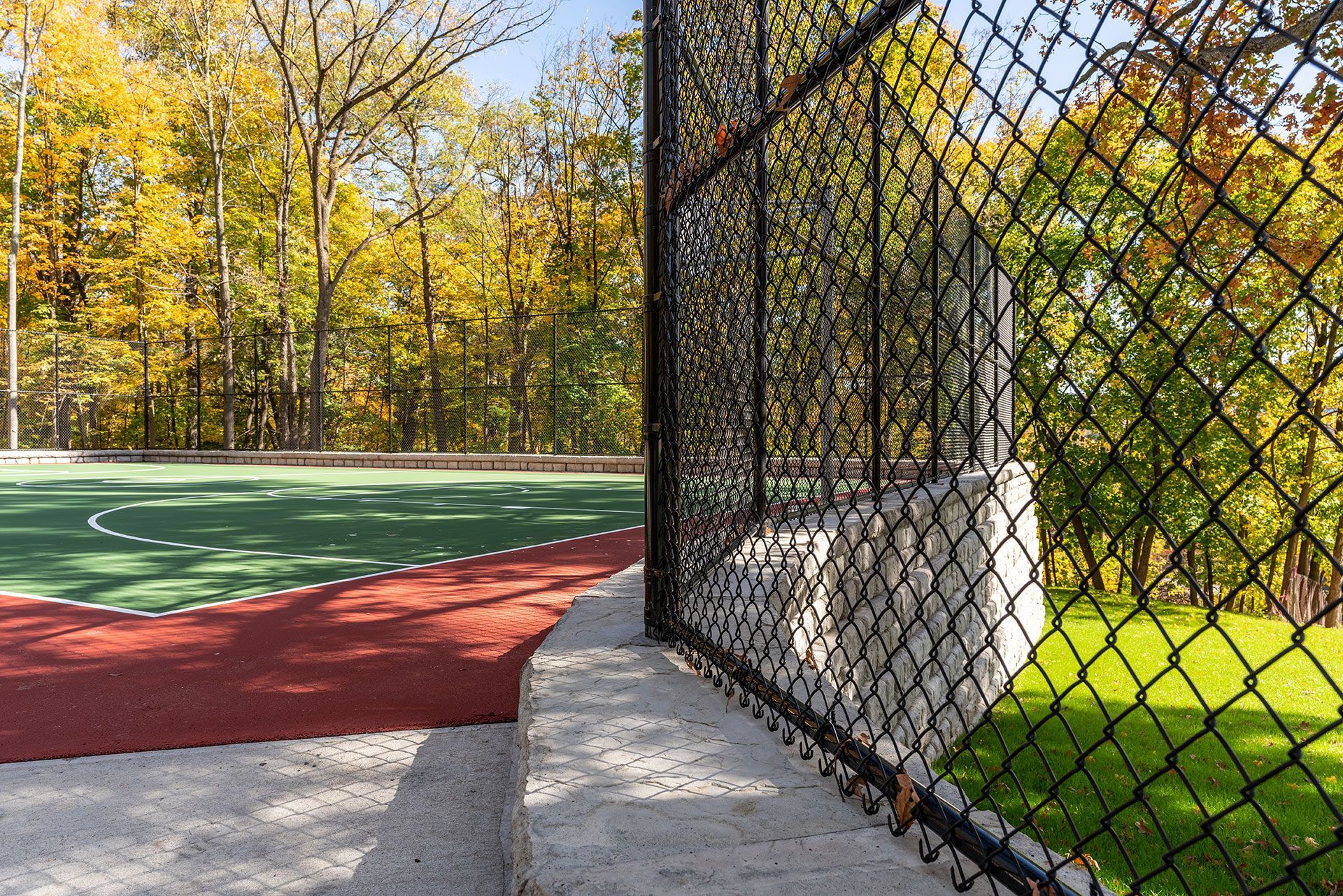 Chain-link fence borders a red and green sports court. Trees with yellow leaves in background.