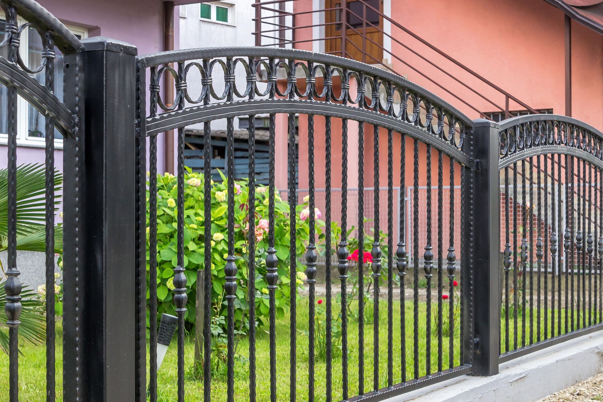 Black wrought iron fence with arched top, surrounding a green lawn with shrubs and a building in the background.