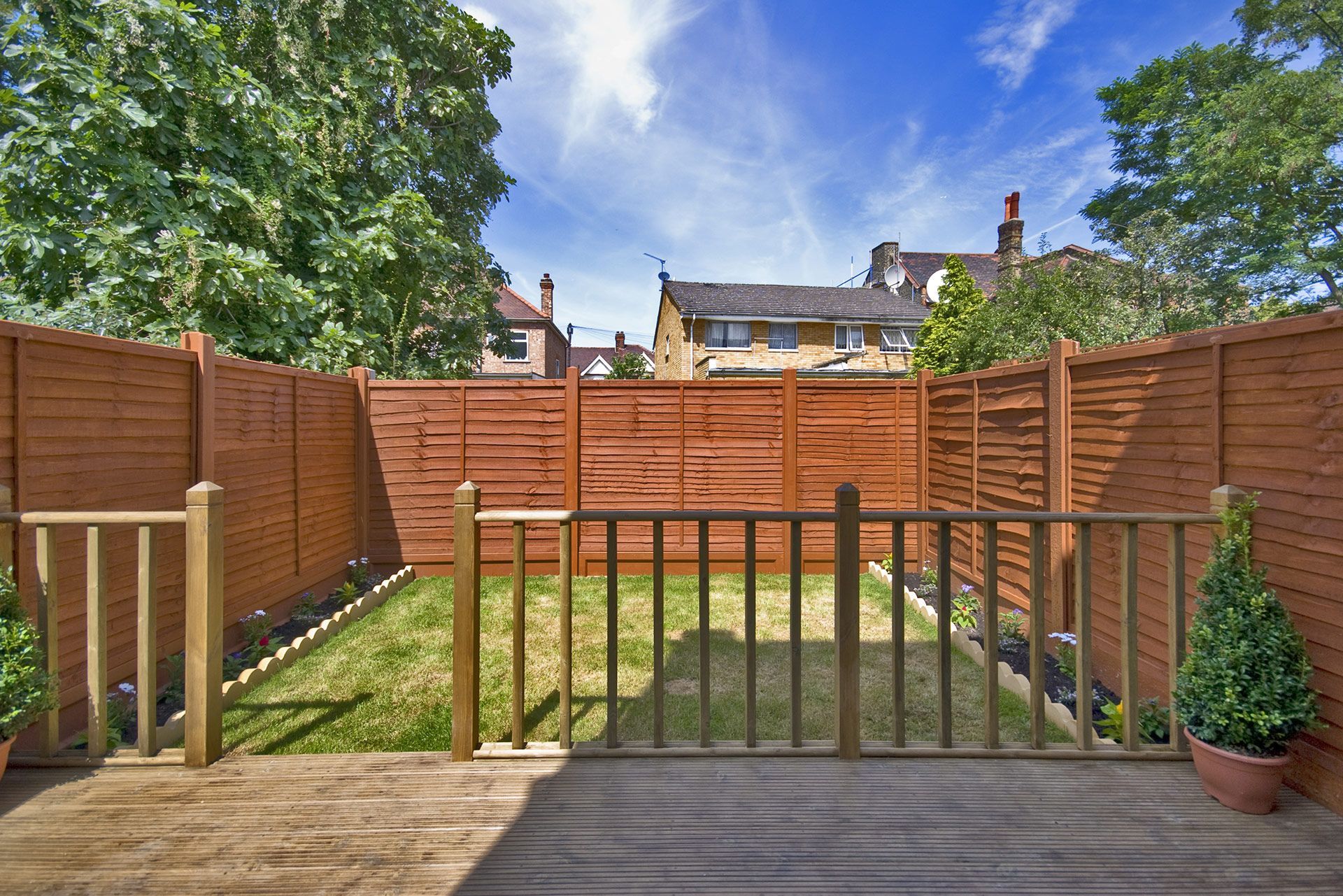 Wooden deck with small lawn bordered by brown fences under a blue sky, houses in the background.