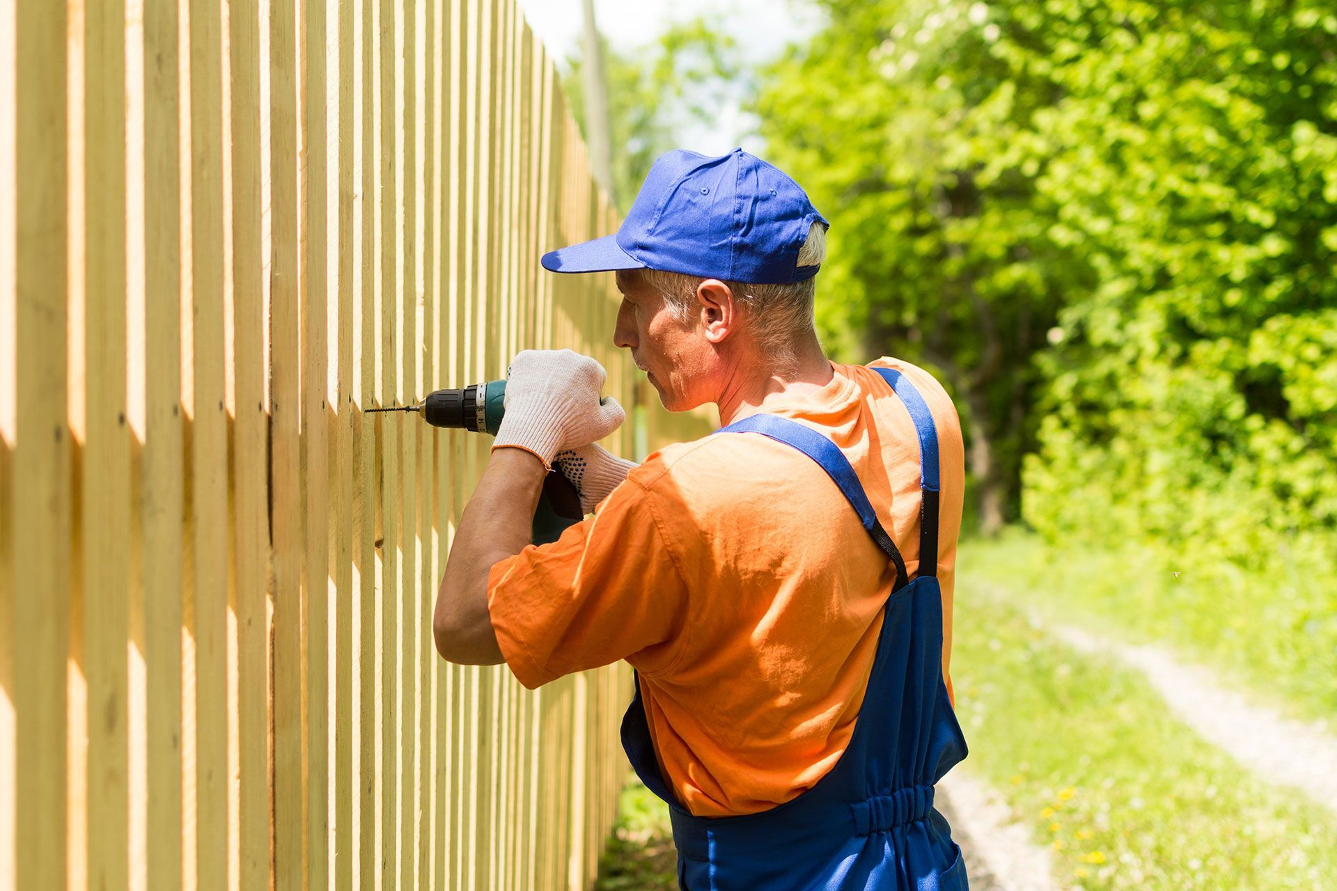 Man in overalls and cap using a drill to install wooden fence panels outdoors.