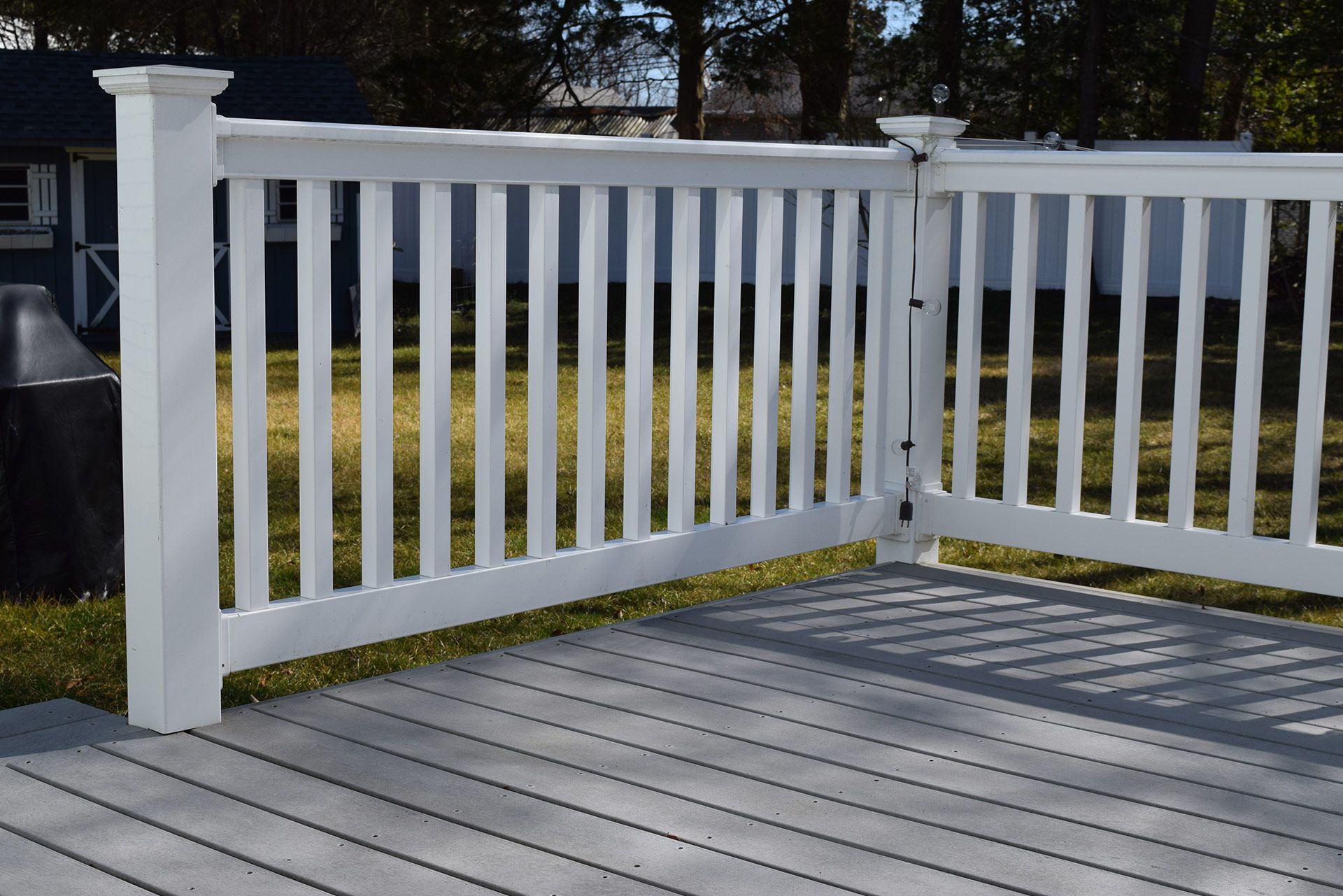 White picket fence on gray deck. Green grass and blue shed in background.
