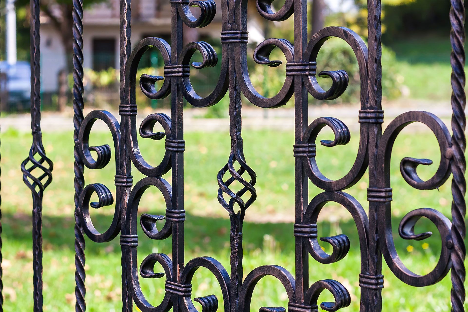 Black wrought iron gate with swirling details, set against a blurred green lawn and building.