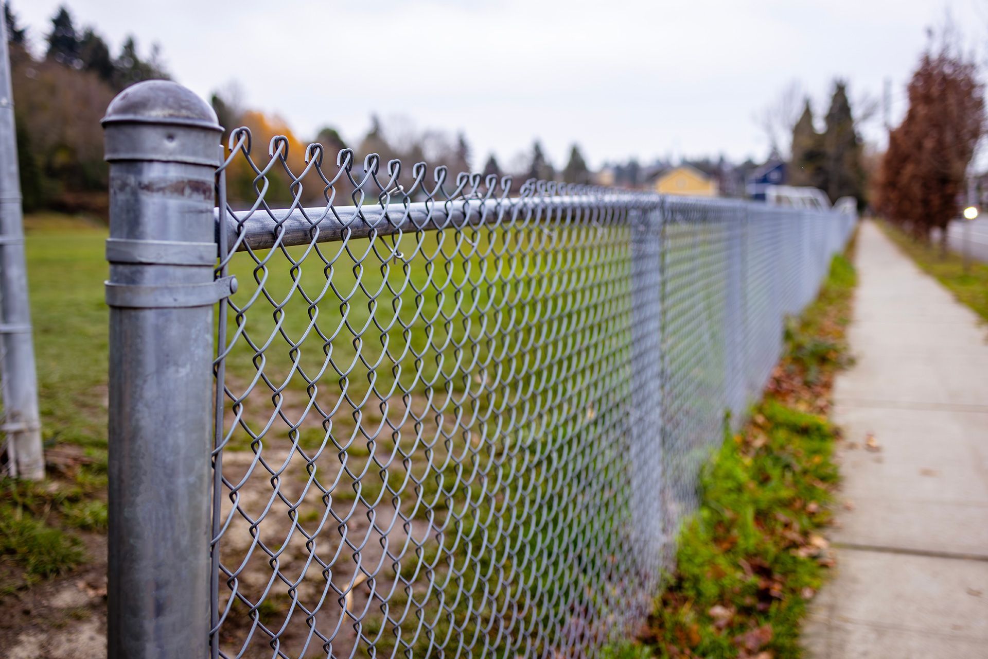 Chain-link fence borders a grassy field next to a sidewalk on a cloudy day.