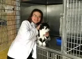 A woman in a lab coat is holding a black and white cat in a cage.