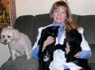 A woman is sitting on a couch holding two cats and a dog.