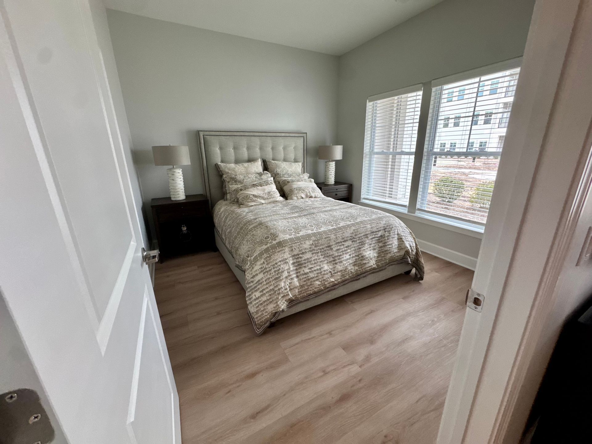 Bright bedroom with a bed, nightstand, and large window blinds over wood flooring.
