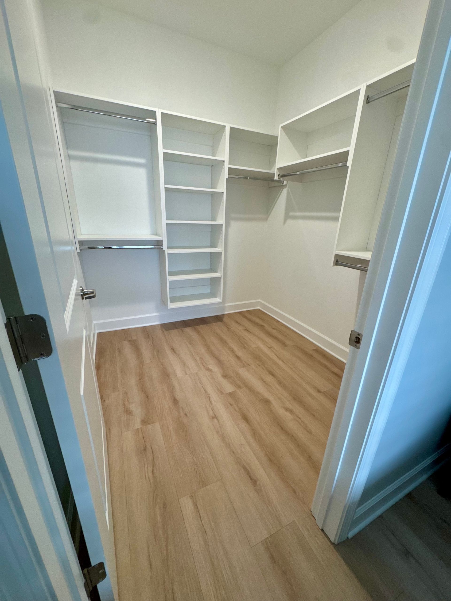 Small empty walk-in closet with white shelves, hanging rods, and light wood flooring.