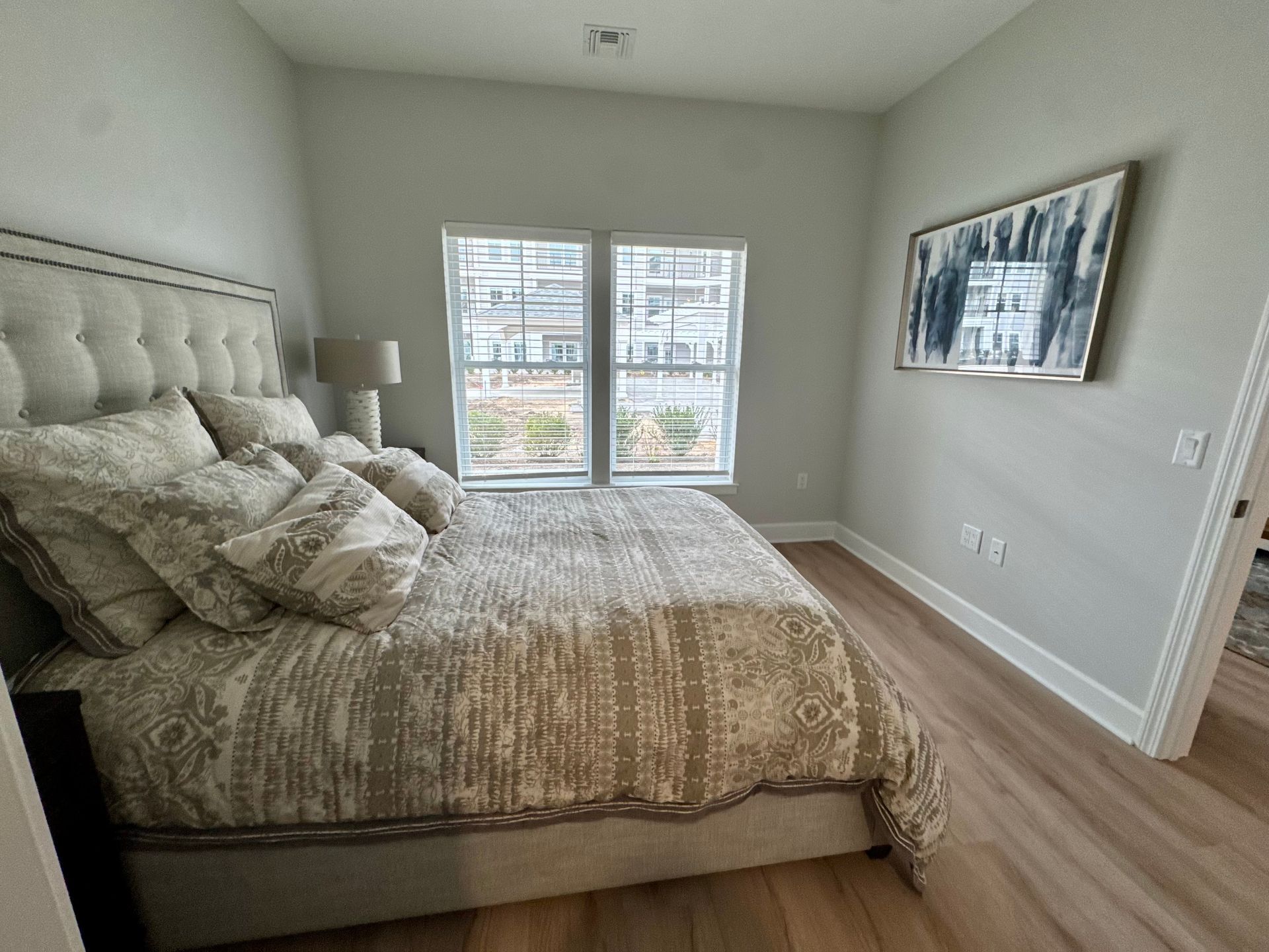 Bright bedroom with tufted bed, patterned bedding, wood floors, and window with city view.