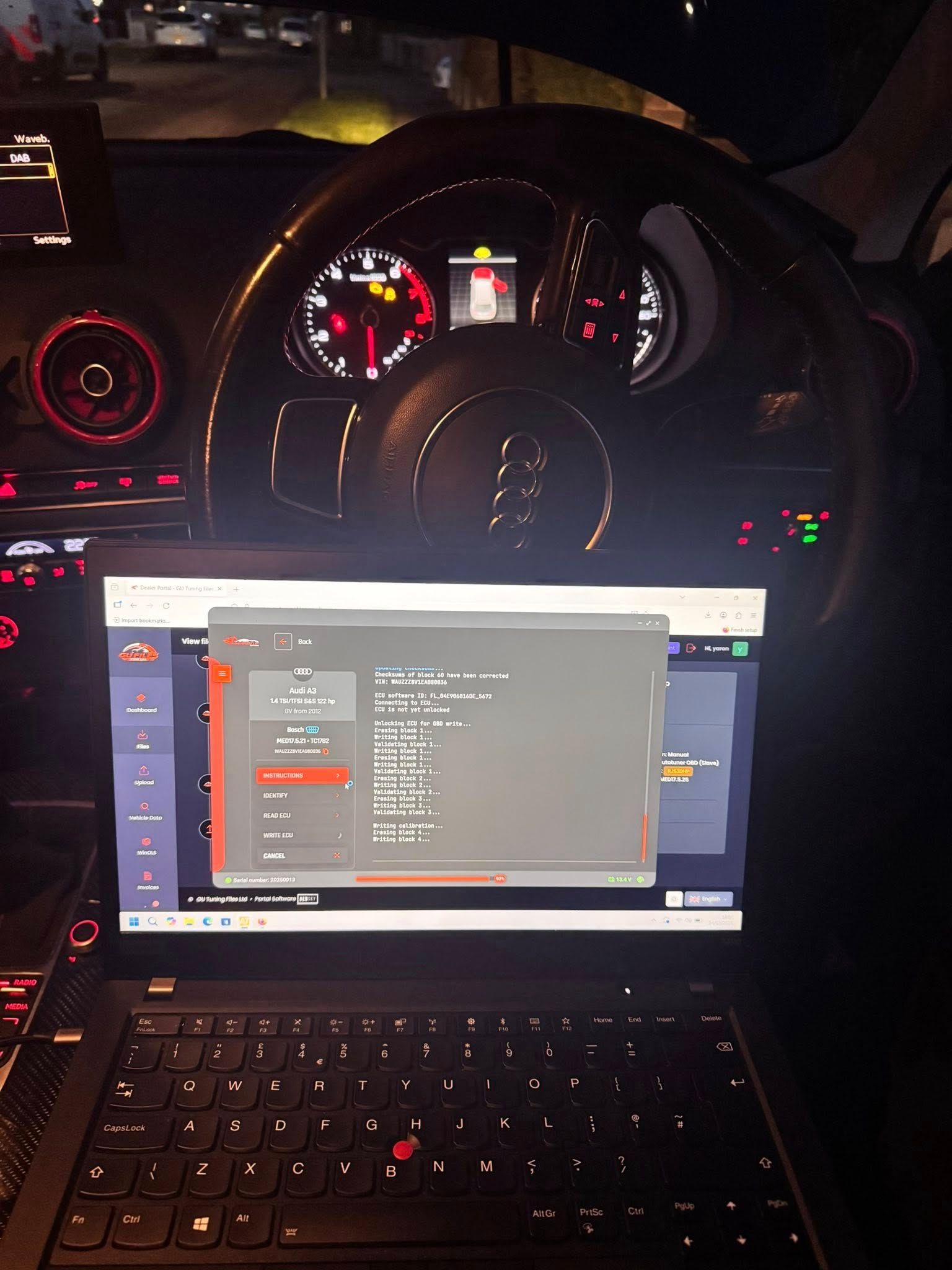 Laptop open in a dark car interior, with a vehicle dashboard glowing red behind it.