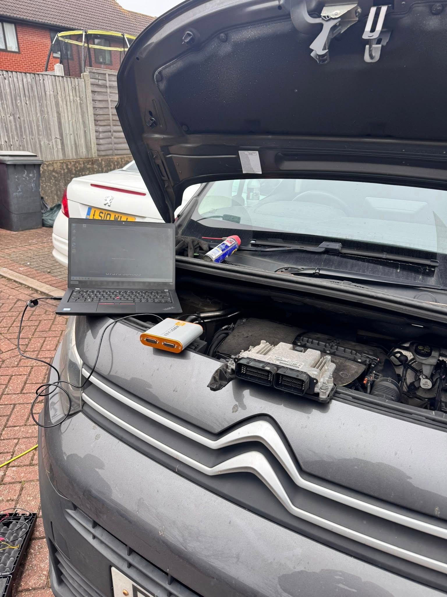Open car hood with a laptop and tools resting on the front bumper in a driveway.