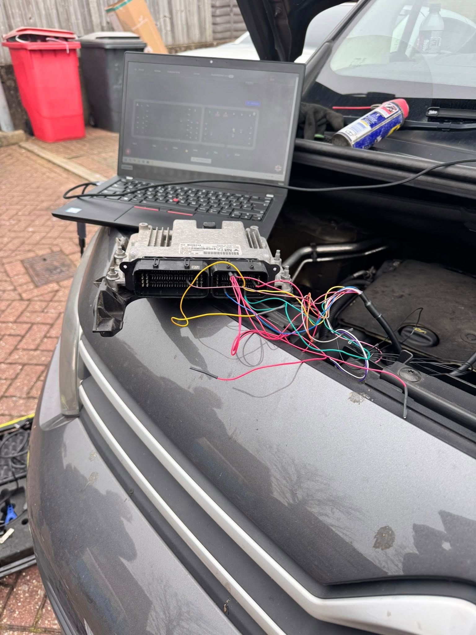 Laptop connected to exposed car wiring on a car hood, with tools and cables in an outdoor driveway