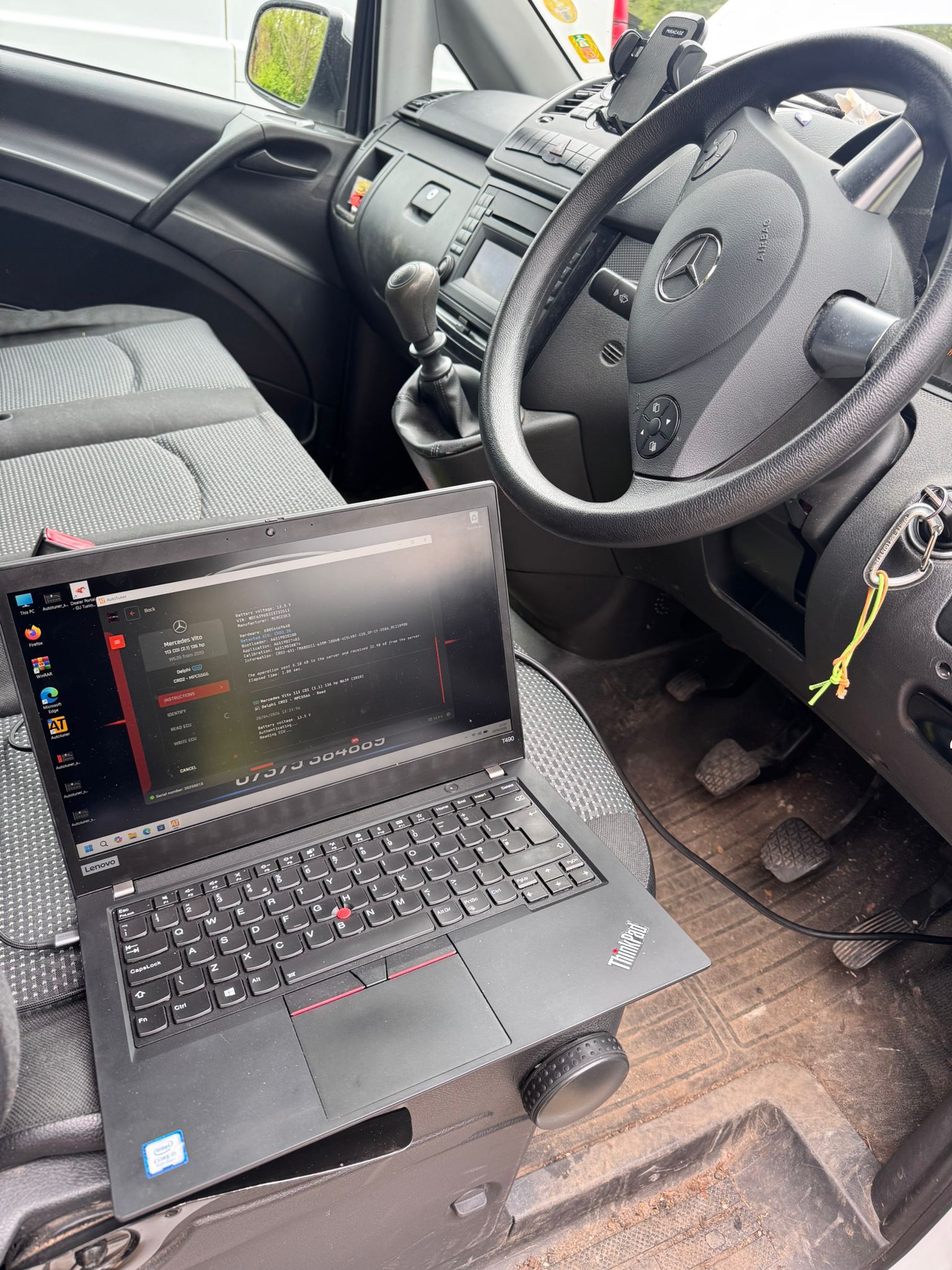 Laptop open on a car passenger seat, showing diagnostics data beside the steering wheel in a parked vehicle.